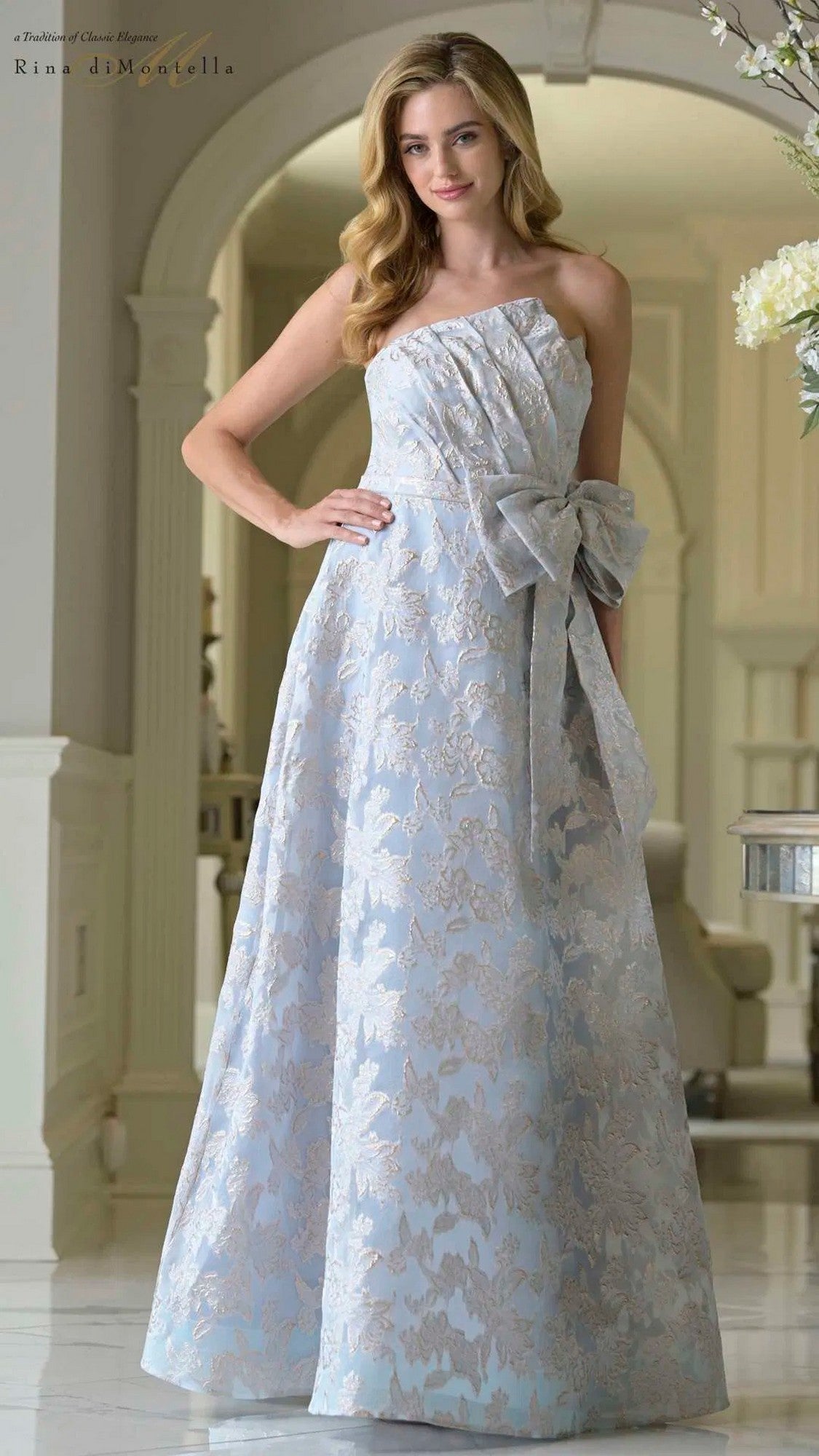 A woman stands indoors in an elegant setting, wearing the Rina Di Montella RD3427—a strapless, pale blue jacquard formal dress featuring a floor-length a-line skirt, floral patterns, and a large waist bow.